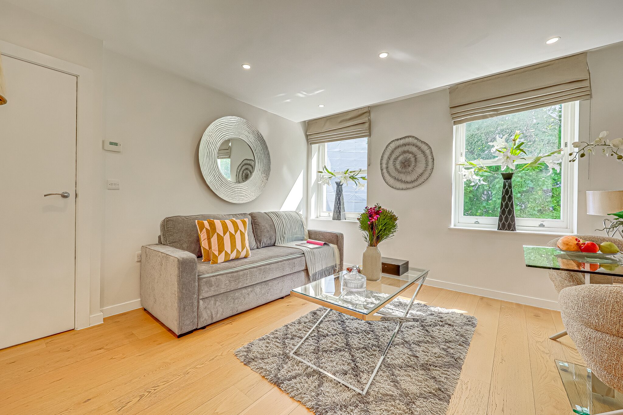 A modern living room with a grey sofa, a glass coffee table, and a large window with a view of greenery.