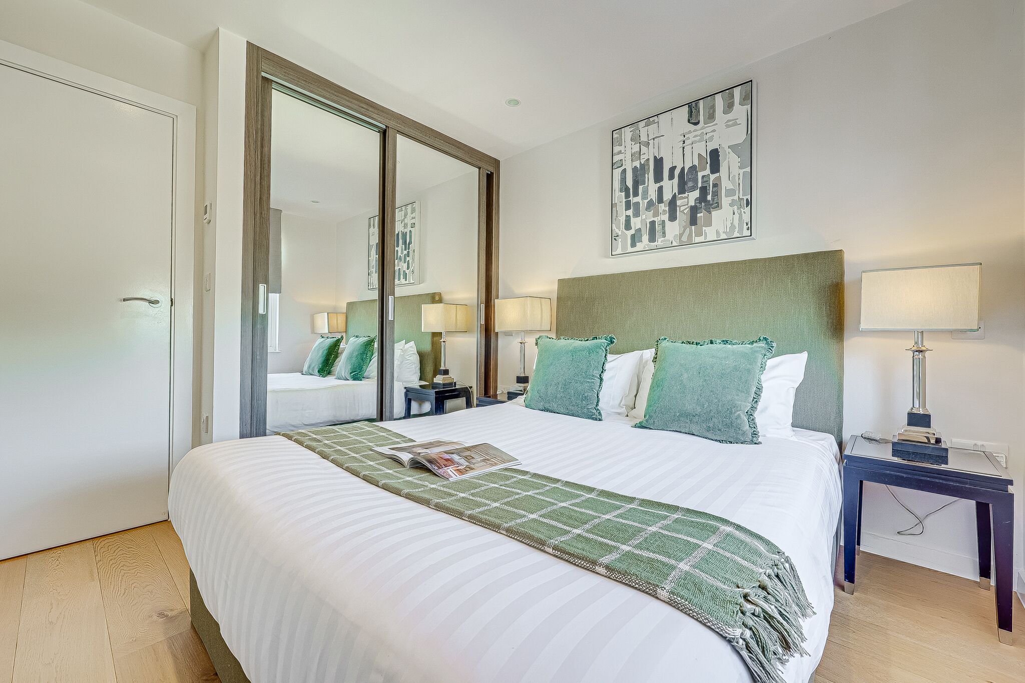 A hotel room with a large bed, a bedside table with a lamp, a mirror, and a wall-mounted artwork.