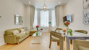 A modern living room with a sofa, a glass coffee table, a dining table with chairs, a TV, and a large window with curtains.