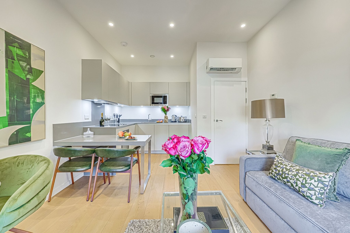 Una sala moderna con un sofá, una mesa de comedor con sillas, una zona de cocina al fondo y un jarrón con flores sobre una mesa auxiliar.