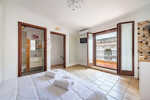Una habitación de hotel con cama, baño, televisión y balcón con mobiliario exterior.