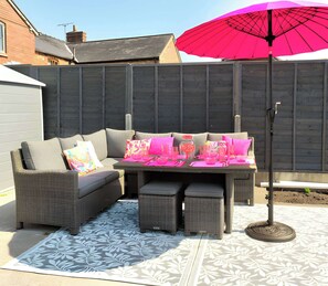 Eine Terrasse mit einer grauen Rattan-Couch, einem Tisch mit rosafarbenen Kissen und einem rosa Sonnenschirm.