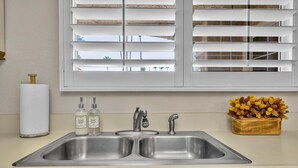 A kitchen with a double sink, a window with blinds, a roll of paper towels, two bottles of soap, and a vase with dried flowers. - Mesquite Breeze - A Ryson Property (Palm Springs)