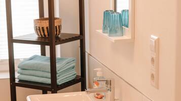 A bathroom with a white sink, a mirror, a shelf with folded towels, and a small potted plant.