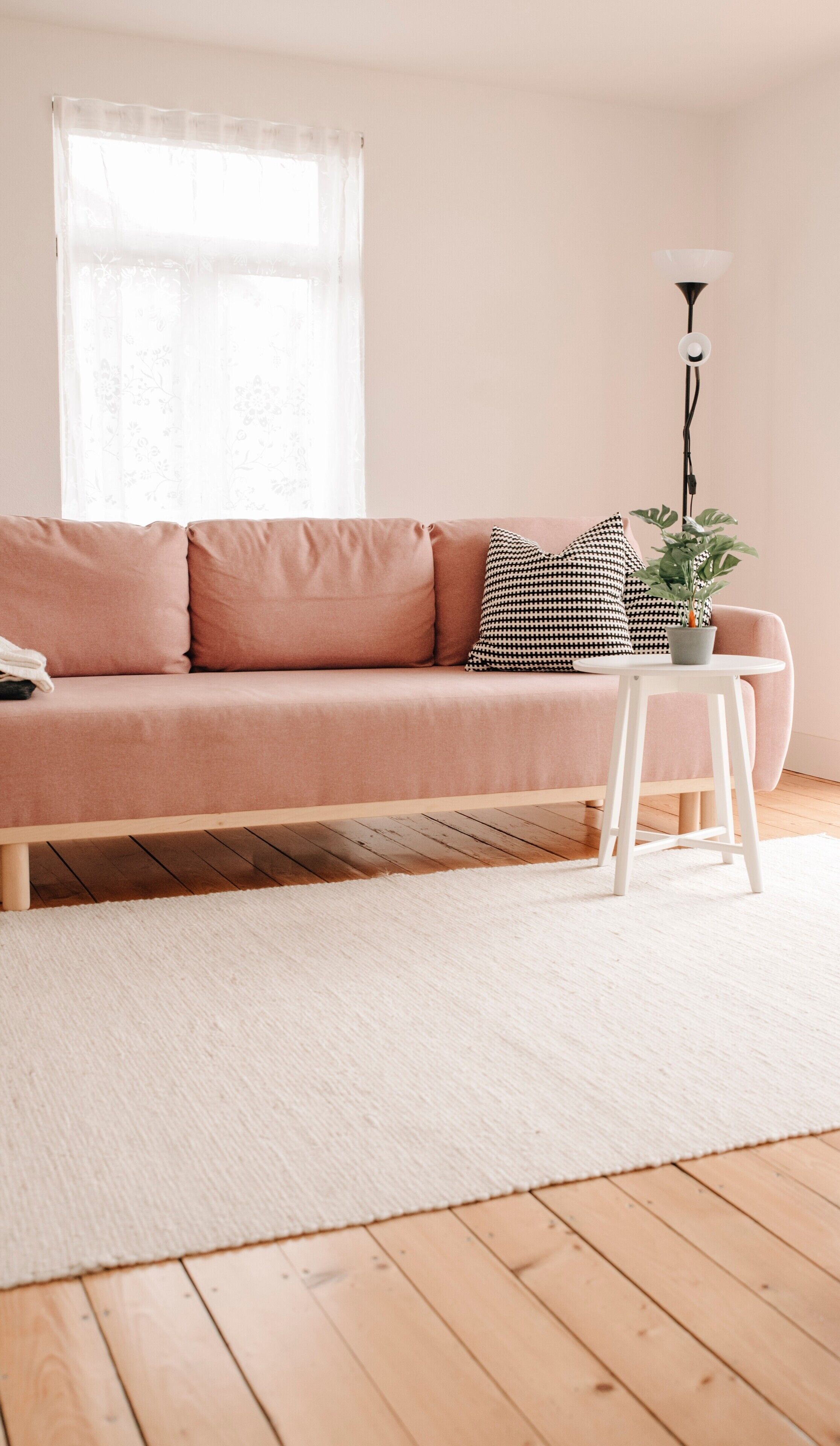 Ein rosafarzenes Sofa mit einem hundezahn-Muster Kissen, ein weißer Nachttisch mit einer Pflanze und eine Stehlampe in einem Wohnzimmer mit Holzboden und einem weißen Teppich.