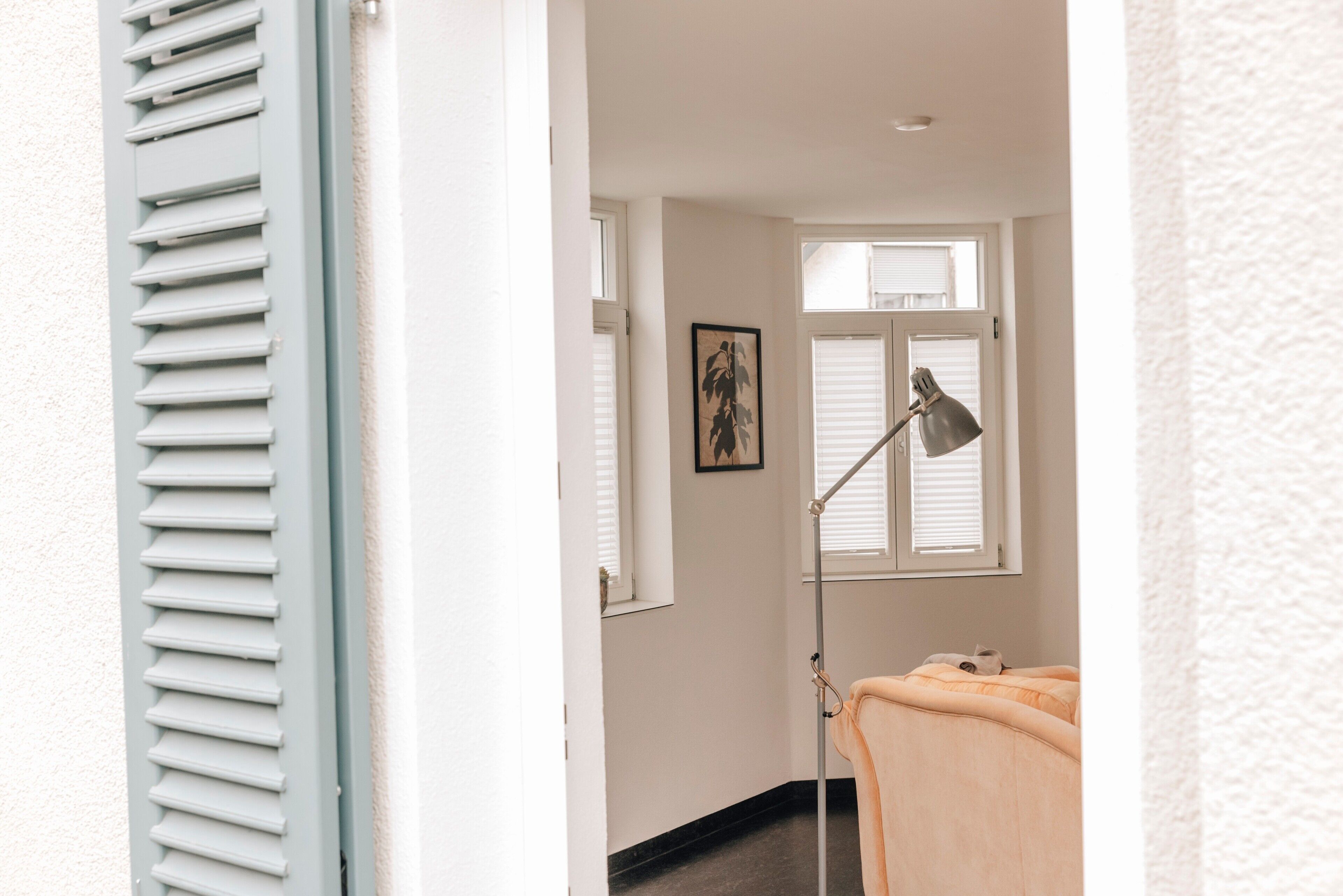 Ein Zimmer mit einem Bett, einer Stehlampe, einem gerahmten Bild an der Wand und einem Fenster mit Jalousien.