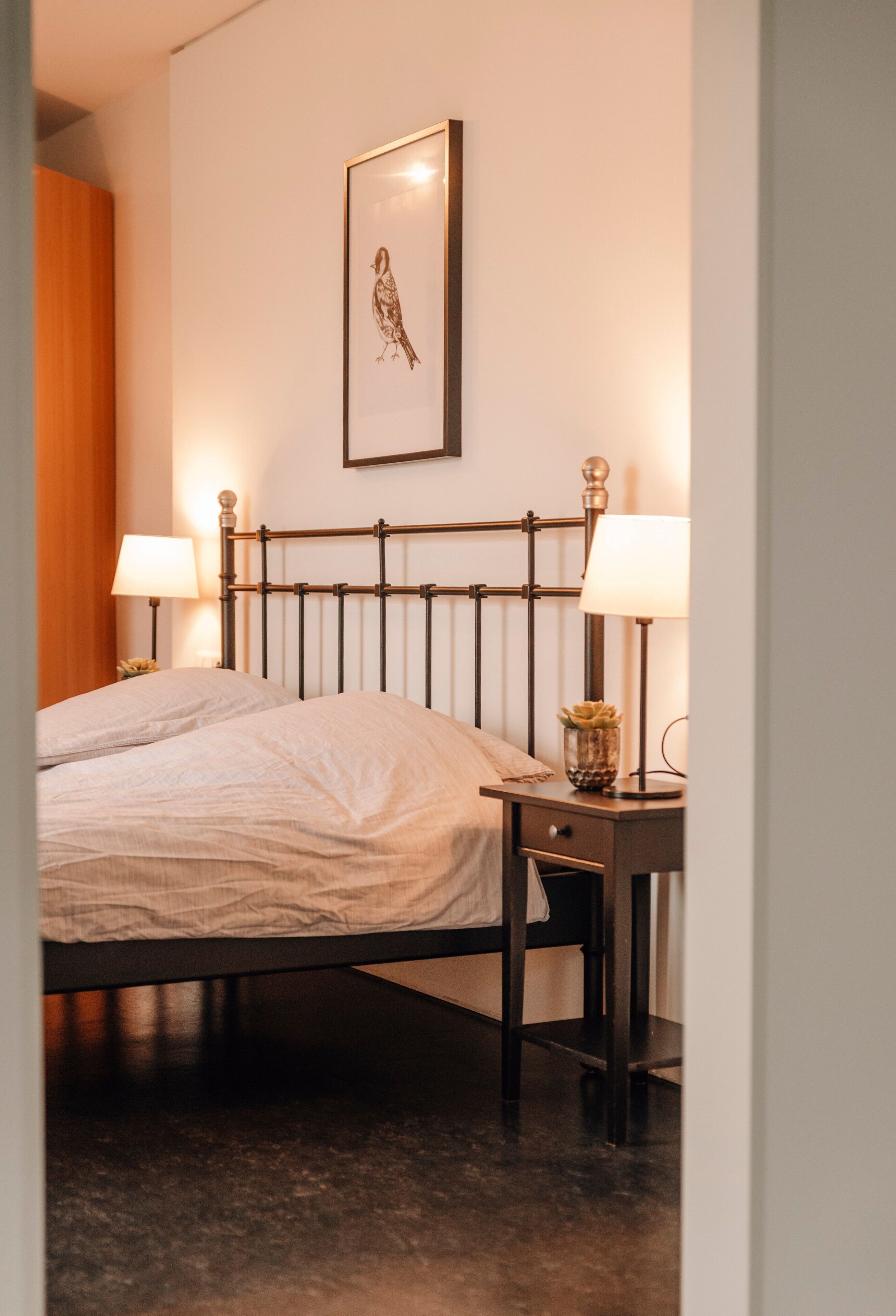 A neatly made bed with a headboard, two bedside lamps, a framed picture on the wall, and a small table with a lamp and a plant.