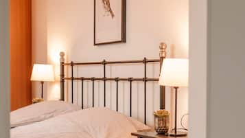 A neatly made bed with a headboard, two bedside lamps, a framed picture on the wall, and a small table with a lamp and a plant.