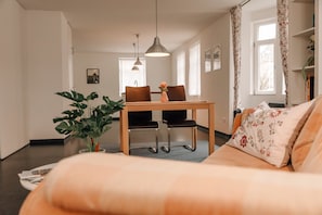Ett modernt vardagsrum med en soffa, ett matbord med stolar, en hängande lampa och en krukväxt.