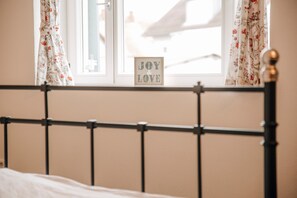 A bed with a metal headboard, a window with floral curtains, and a sign that says 'JOY & LOVE' on the window sill.