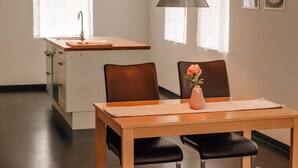 A dining area with a wooden table, two chairs, a vase with a flower, pendant lights, a kitchen in the background, and a window with curtains. - Apartment 'Ferienwohnung Ambiente Am Federsee' with Wi-Fi (Bad Buchau)