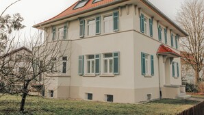 Un bâtiment à deux étages, avec un toit rouge, de nombreuses fenêtres dotées de volets verts et un petit porche à l’avant.