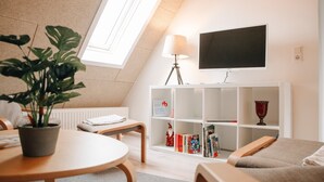 Un salon moderne avec un plafond en pente, un téléviseur fixé au mur, une bibliothèque, une plante en pot et un coin salon confortable.