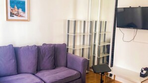 Un salon avec un canapé violet, un pouf gris, un téléviseur fixé au mur et une étagère.