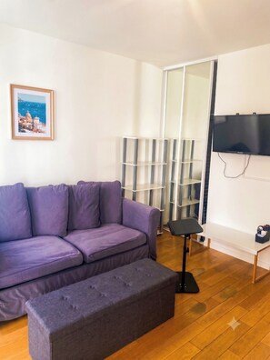 Ein Wohnzimmer mit einem violetten Sofa, einem grauen Pouf, einem an der Wand montierten Fernseher und einer Regaleinheit.