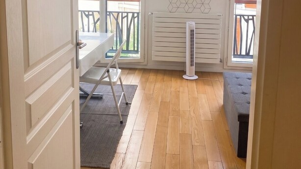 Une pièce avec un plancher en bois, un radiateur blanc, une table avec des chaises et une fenêtre donnant sur des immeubles.