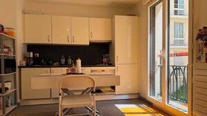 Une cuisine moderne avec des meubles blancs, un coin repas avec une chaise et une grande fenêtre donnant sur l’extérieur.