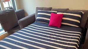 Un lit recouvert d’une couvre-lit à rayures, un seul coussin aux couleurs vives et une chaise dans une pièce dont le sol est en bois.