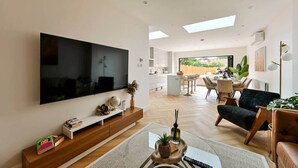 A modern living room with a large flat-screen TV, a dining area, and a kitchen visible through an open door. - 5BR Family Home | Garden | AC | Near Wembley (Greenford)
