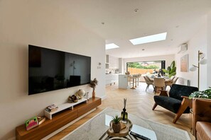 A modern living room with a large flat-screen TV, a dining area, and a kitchen visible through an open door. - 5BR Family Home | Garden | AC | Near Wembley (Greenford)