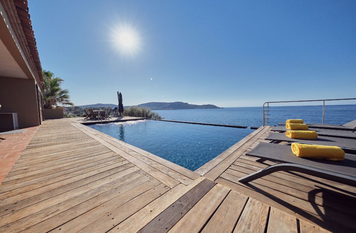 Una terrassa de fusta amb piscina, cadiretes de platja amb tovalloles i vista al mar i les colines llunyanes.