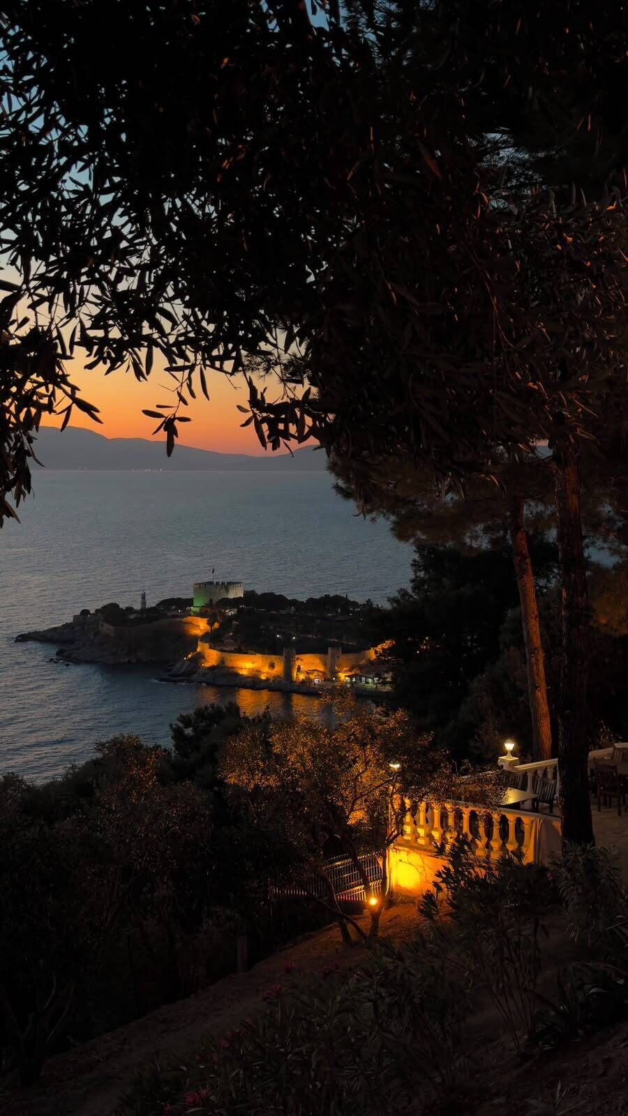 A coastal view with a building illuminated by warm lights, a body of water, and a tree in the foreground.