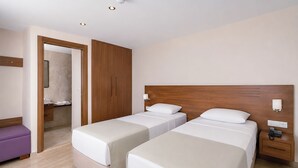 Habitación de hotel con dos camas, un armario de madera, un baño visible a través de una puerta abierta y un pequeño banco morado.