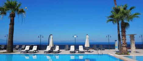 Zona de piscina con sillones de descanso y sombrillas, cielo azul despejado y vista al mar.