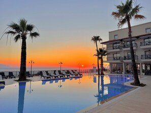 A poolside area with lounge chairs, palm trees, and a building with balconies. - ALTAMARE HOTEL (Kusadasi)