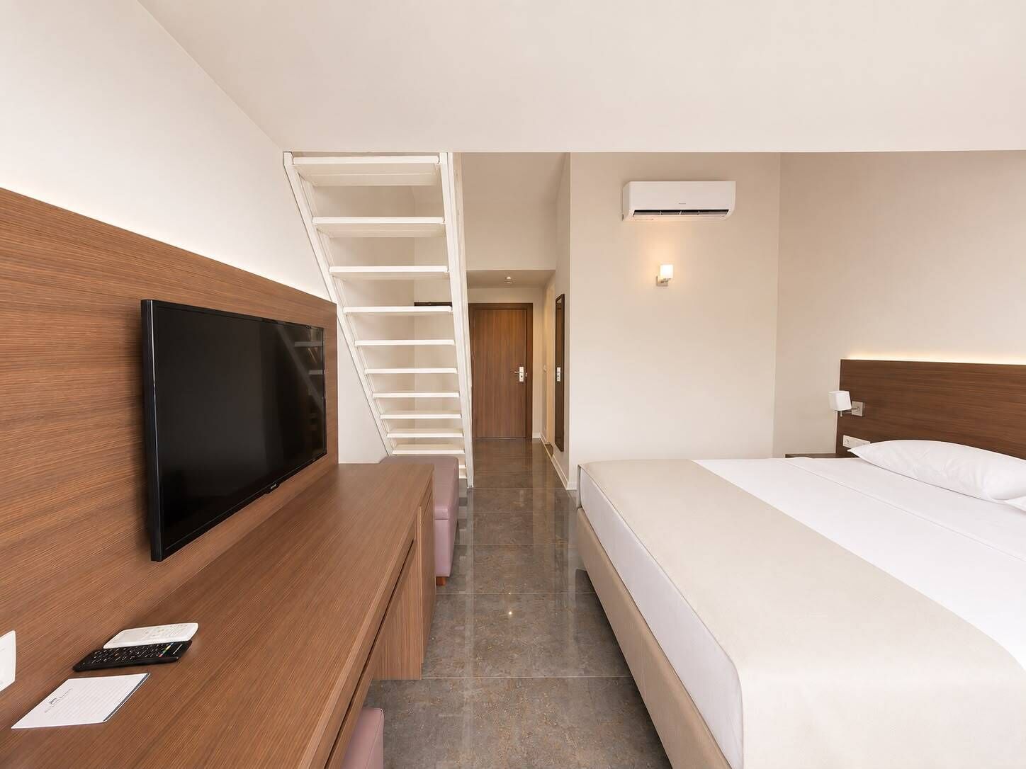 Una habitación de hotel con cama, televisión sobre un escritorio de madera, una escalera y un equipo de aire acondicionado.