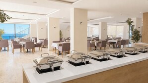 Un área de comedor moderna con varias mesas y sillas, un mostrador de buffet con calentadores de alimentos, y grandes ventanales con vista al mar.