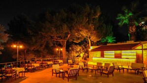 Un área de estar al aire libre con mesas y sillas, un edificio de piedra con un bar, y árboles al fondo.