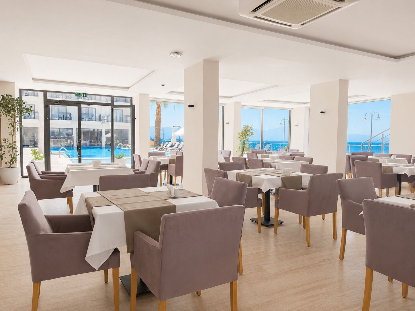 A modern dining area with tables and chairs, large windows with a view of a pool and the sea, and a ceiling with recessed lighting.
