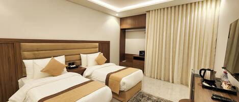 A hotel room with two beds, a desk, a chair, a coffee maker, and a large window with curtains.