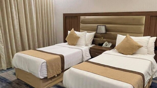 Habitación de hotel con dos camas, cabecero de madera, mesita de noche con lámpara y ventana con cortinas.