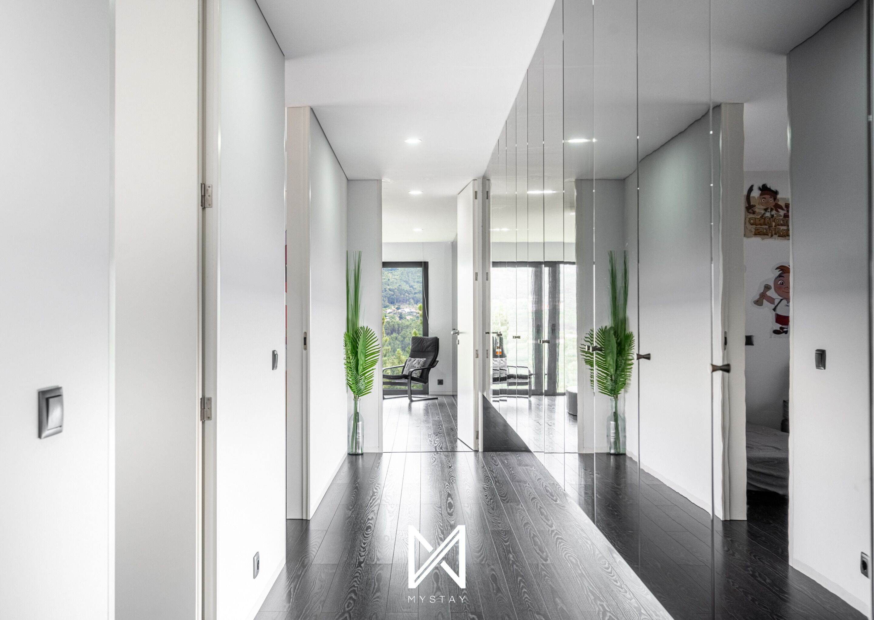 A modern hallway with a wooden floor, white walls, and glass doors leading to a balcony with a view.