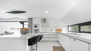 A modern kitchen with white cabinetry, a central island with bar stools, a built-in oven, and a large window providing natural light. - MyStay - Varanda de Basto (Canedo de Basto)