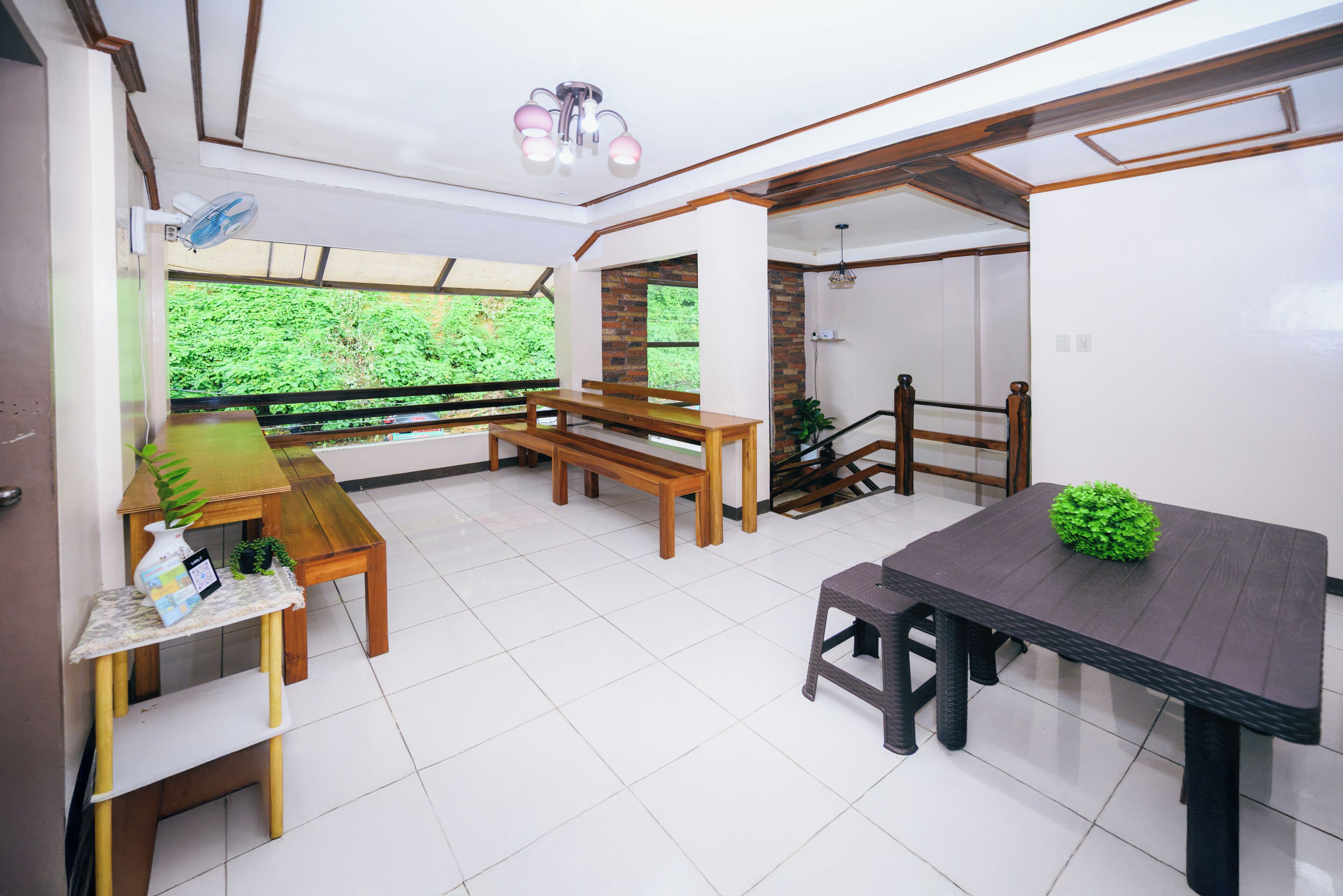 A room with a wooden table, benches, a dark table, and a view of greenery outside.