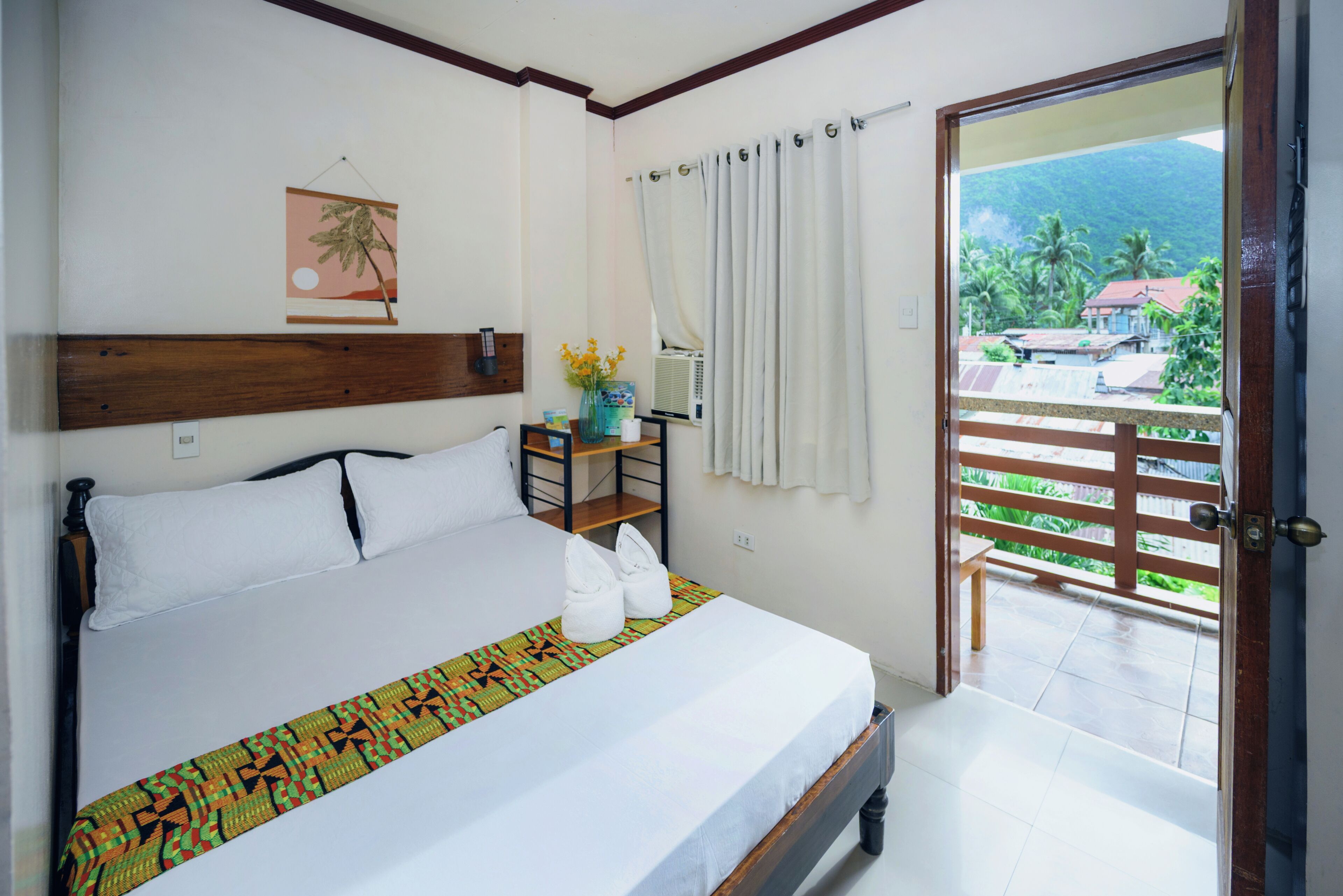 A hotel room with a bed, a small table, a vase with flowers, a window with curtains, and a view of the outside.