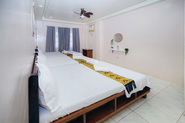 Habitación de hotel con dos camas individuales, un ventilador de techo, una ventana con cortinas, una mesita con un jarrón y una estantería con un espejo y una planta.