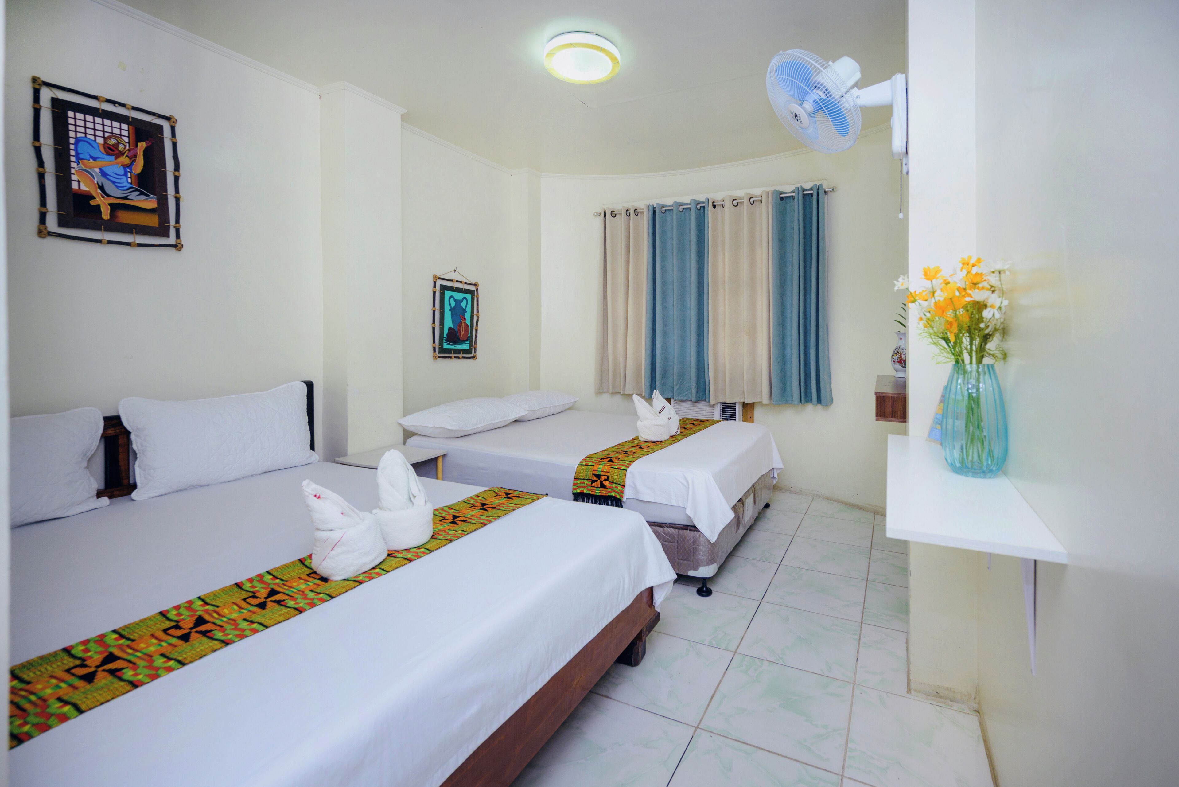Habitación de hotel con dos camas, ventilador de techo, ventana con cortinas y un florero con flores sobre una mesita de noche.
