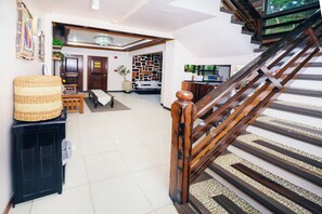 Una escalera con barandillas de madera que conduce a un piso superior, un electrodoméstico negro a la izquierda y un papel tapiz con diseño en el fondo.