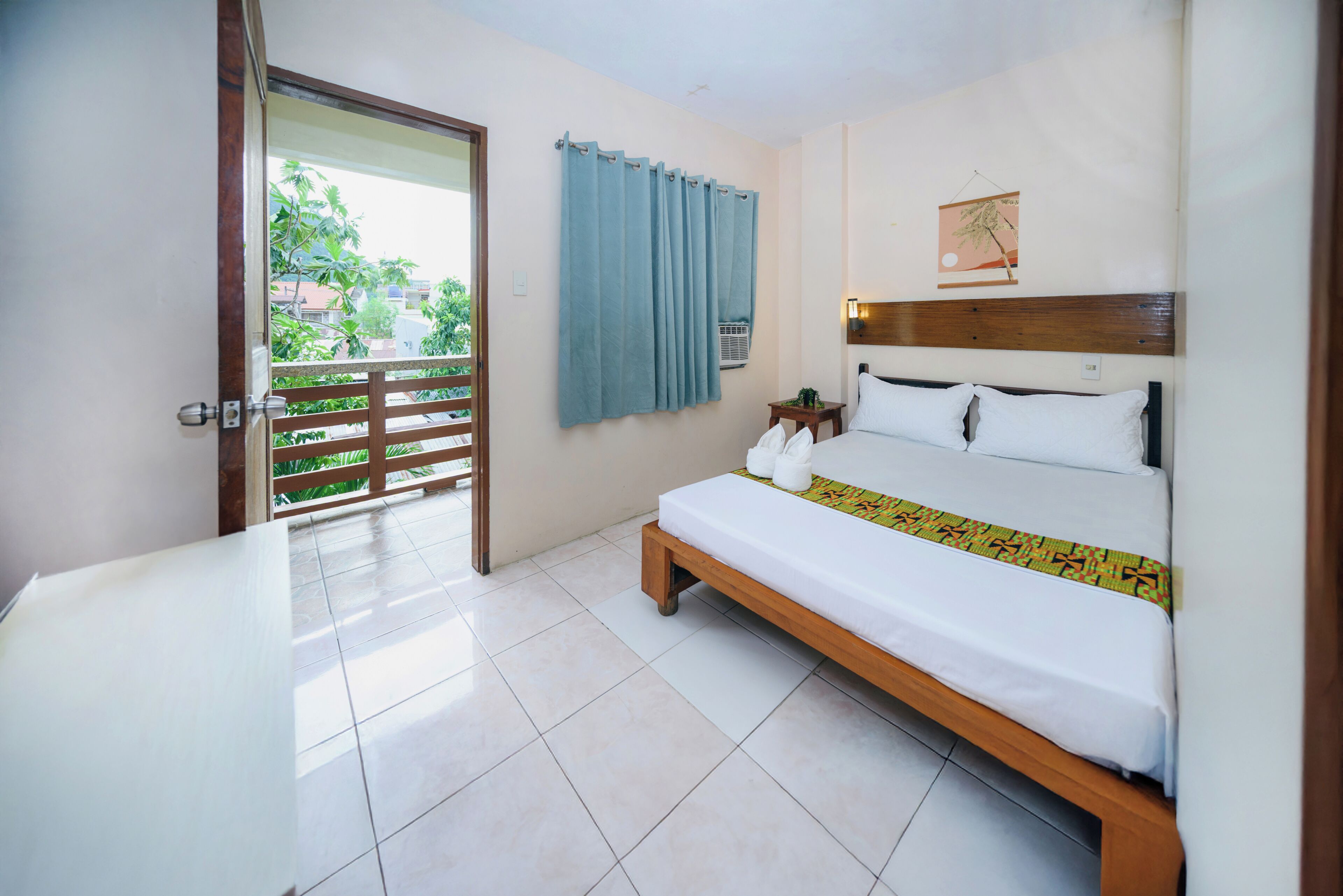 A hotel room with a bed, a small table, a wall-mounted air conditioner, and a view of greenery outside.