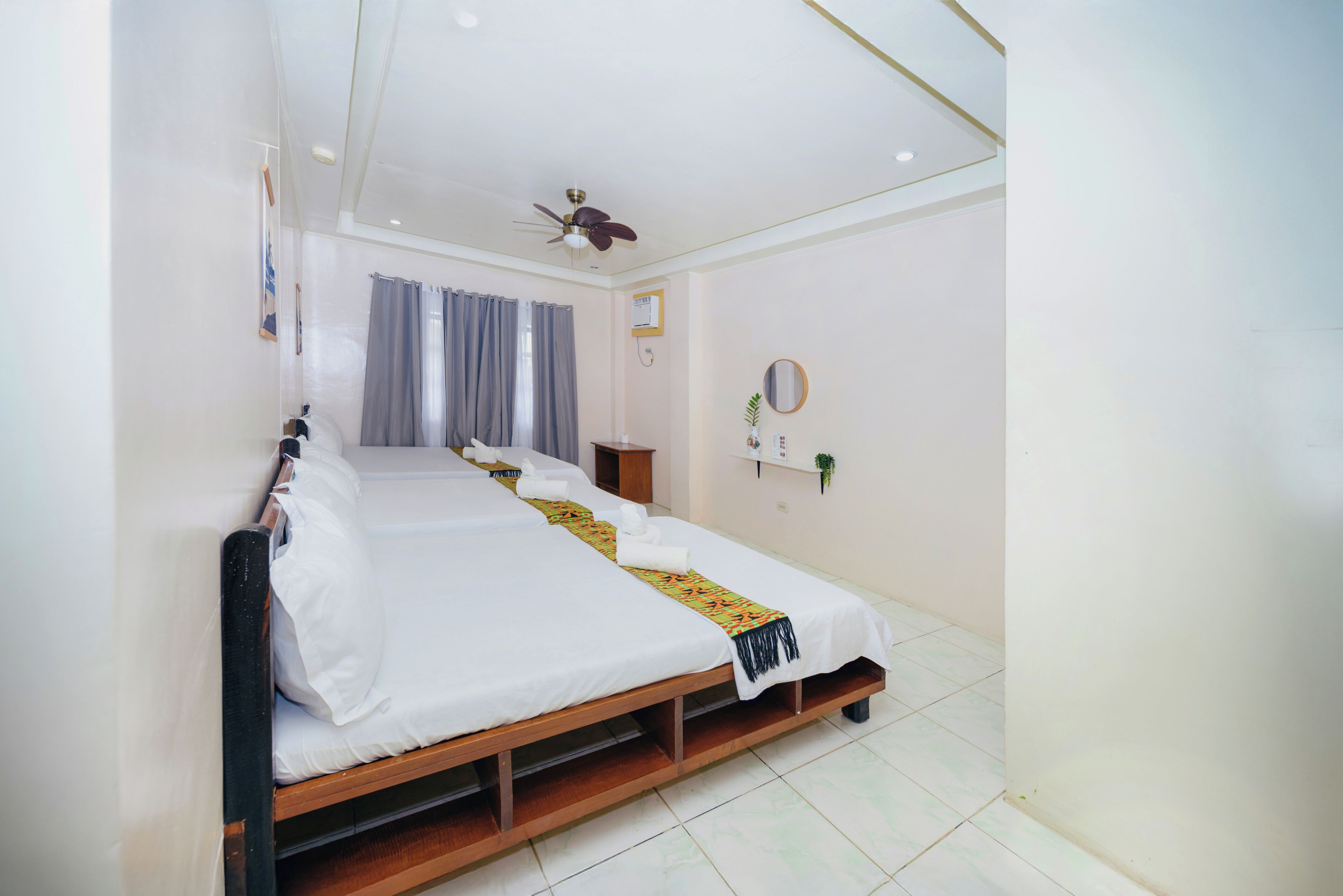 A hotel room with a single bed, a ceiling fan, a small bedside table, a mirror, and a window with curtains.