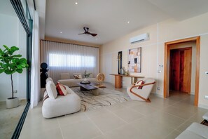A modern living room with a sofa, armchairs, a coffee table, and a floor lamp. There is a potted plant, a ceiling fan, and an air conditioning unit. - Beachfront 2BR Condo w/ Balcony • Sol de Arena E4 (Las Terrenas)