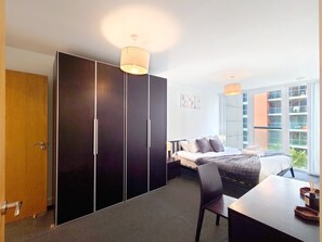A hotel room with a bed, a desk, a wardrobe, and a window with a view. - Deluxe Two Bed Fully Serviced Apartment in ExCeL (London)