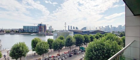 A view of a riverside area with a city skyline, trees, and parked vehicles.