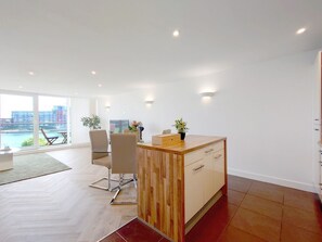 A modern living room with a wooden desk, a chair, and a view of a body of water through large windows. - Deluxe Two Bed Fully Serviced Apartment in ExCeL (London)