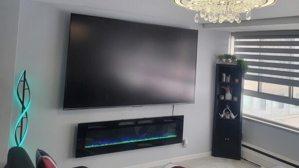 A modern living room with a flat-screen TV, a decorative chandelier, a black entertainment unit, and a window with blinds. - The River Condos - Detroit, Michigan (Detroit)