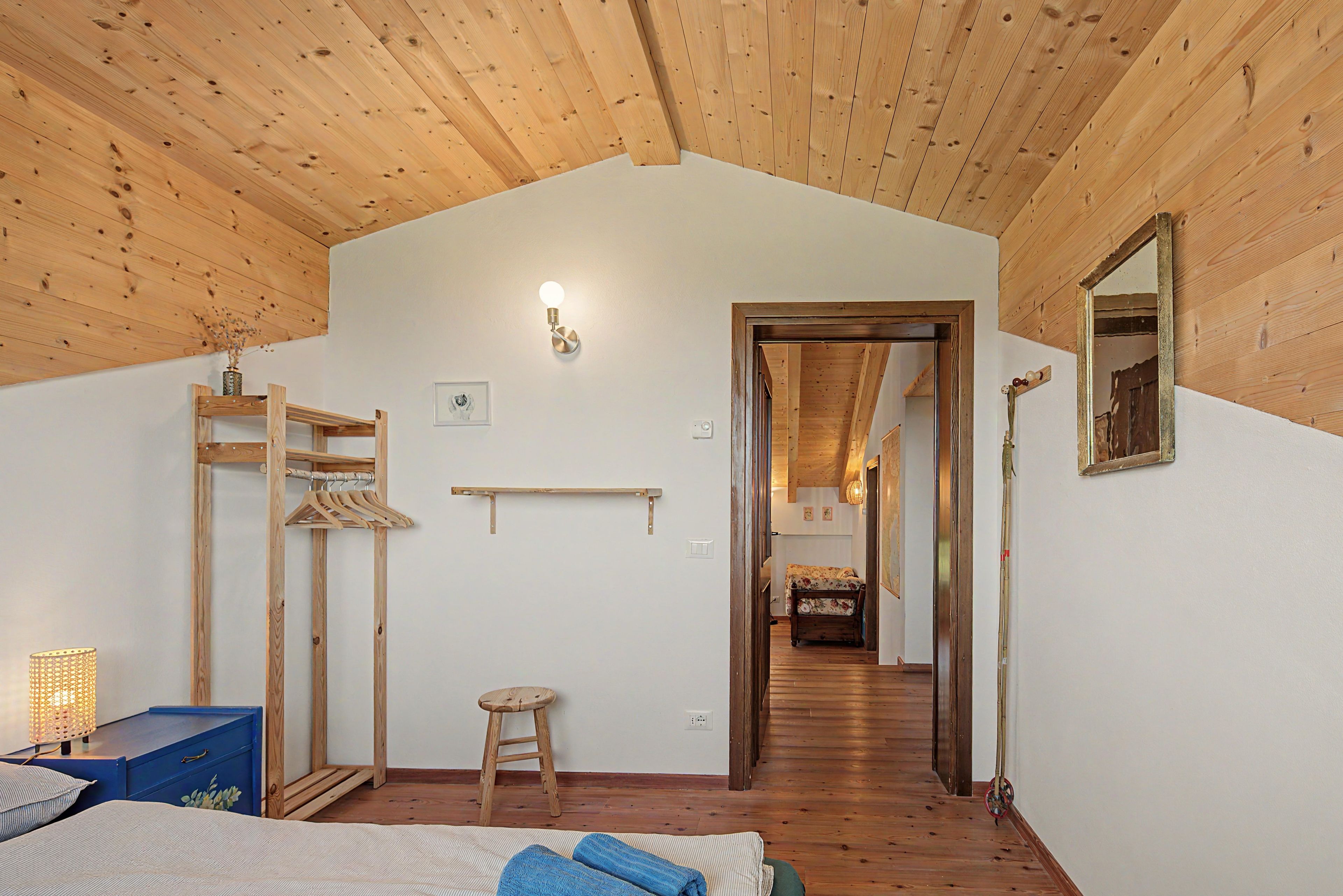 A bedroom with a wooden ceiling, a bed, a hanging clothes rack, a stool, and a hallway leading to another room.